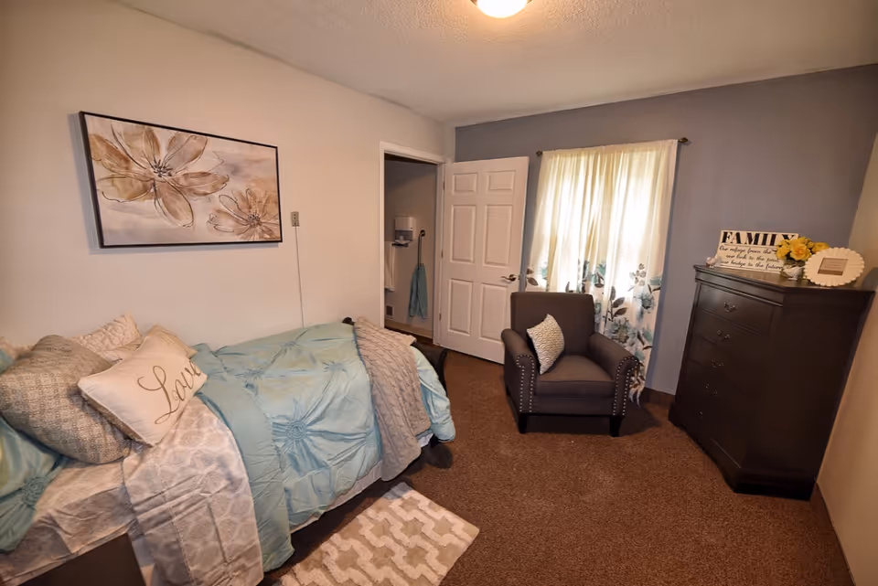 A cozy bedroom with a bed covered in light blue bedding and multiple pillows, including one with the word 'Love'. There is a gray armchair with a decorative pillow near a window with floral curtains. A dark wooden dresser with decorative items, including a sign that says 'FAMILY', is against the wall. A framed floral artwork hangs above the bed, and the room has beige carpet and neutral-colored walls.