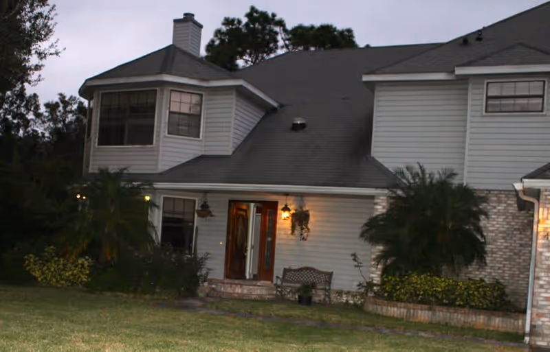 Exterior view of a two-story residential house at dusk with lit porch lights, a wooden front door, windows, and surrounding greenery including bushes and trees.