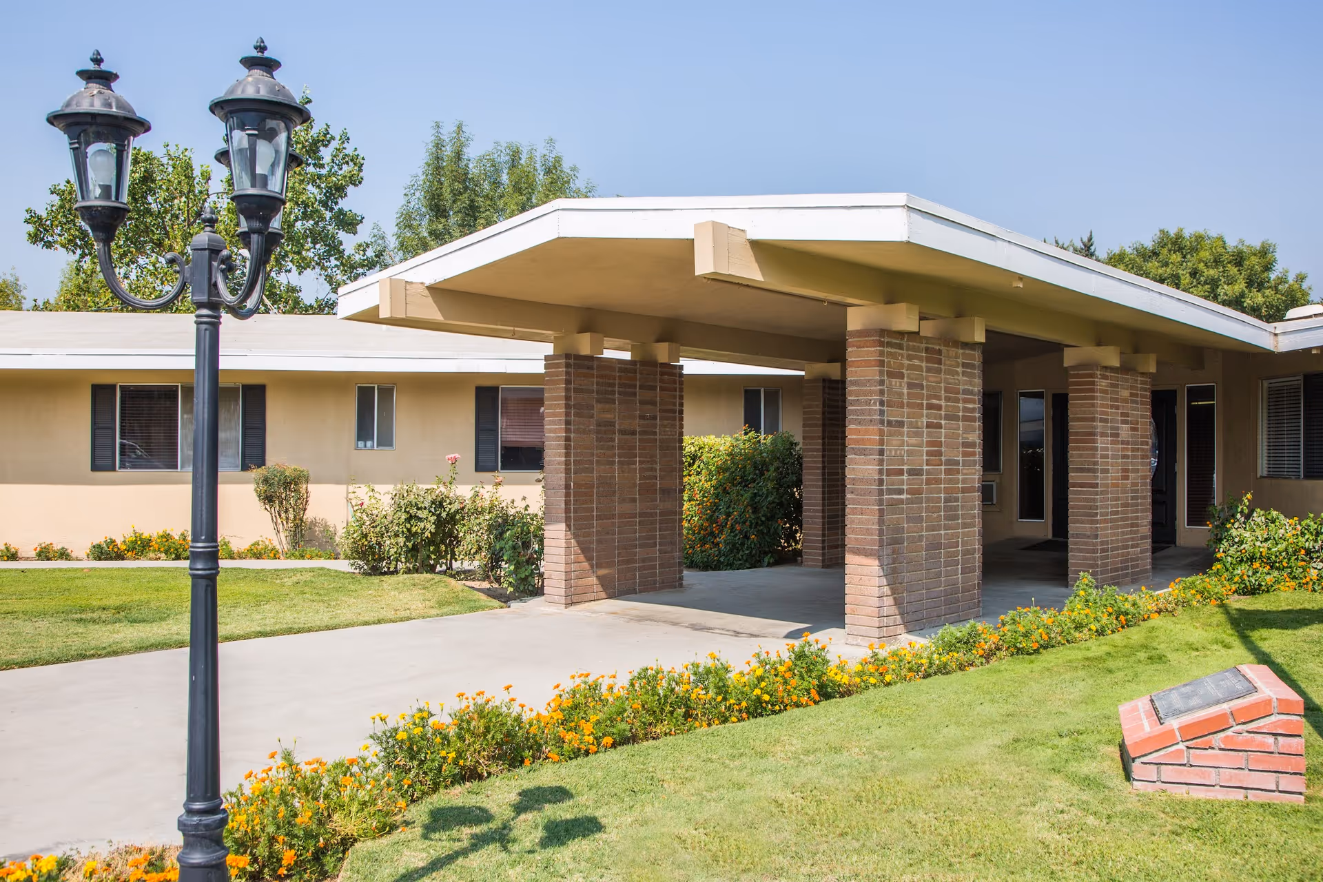 Exterior view of Kingsburg Care Center showing a covered entrance supported by brick columns, a well-maintained lawn with flower beds, a black vintage-style street lamp, and a clear blue sky.