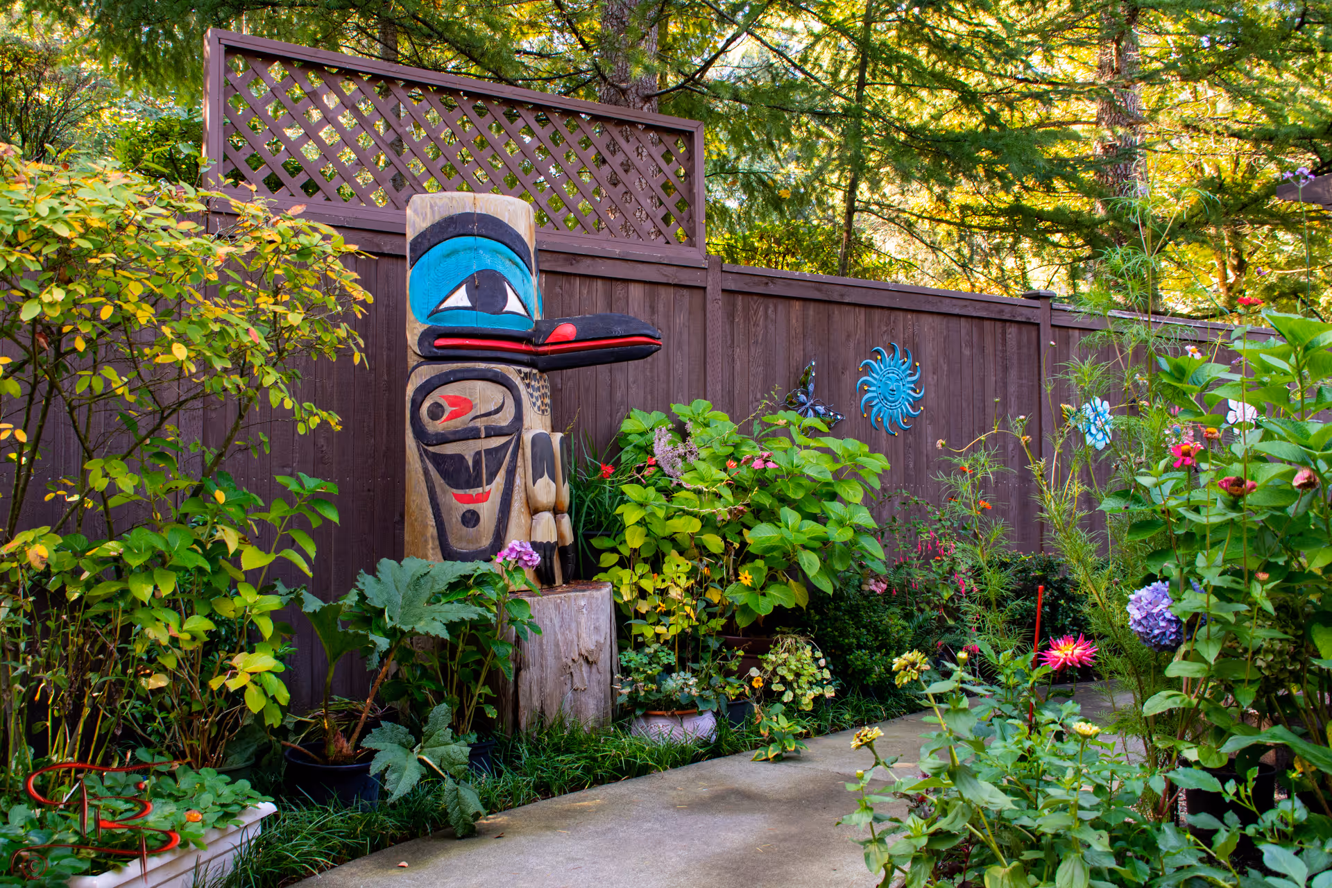 A vibrant garden area with various green plants and colorful flowers along a concrete pathway. A large wooden totem pole with blue, black, and red painted designs stands prominently near a brown wooden fence decorated with blue and black metal sun and flower ornaments. Tall trees and greenery are visible in the background.
