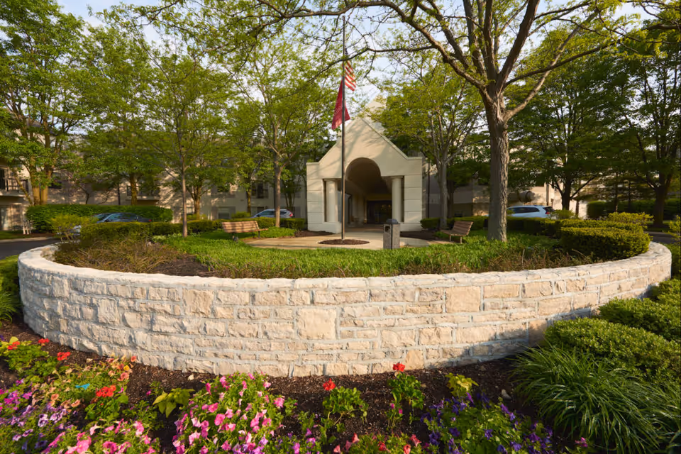 Entrance to a senior living facility with a circular stone wall surrounding a landscaped garden area with flowers and greenery. Two benches are placed inside the circular area near a flagpole with an American flag and another flag. Trees surround the entrance, and cars are parked in the background.