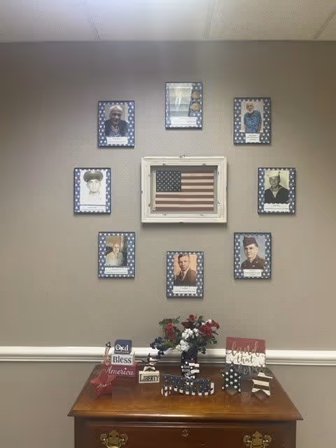 A wall display featuring a framed American flag in the center surrounded by eight framed portraits of veterans, each bordered with a blue star pattern. Below the display is a wooden table with patriotic decorations including flowers, a small American flag, and signs with phrases like 'God Bless America' and 'family first'.