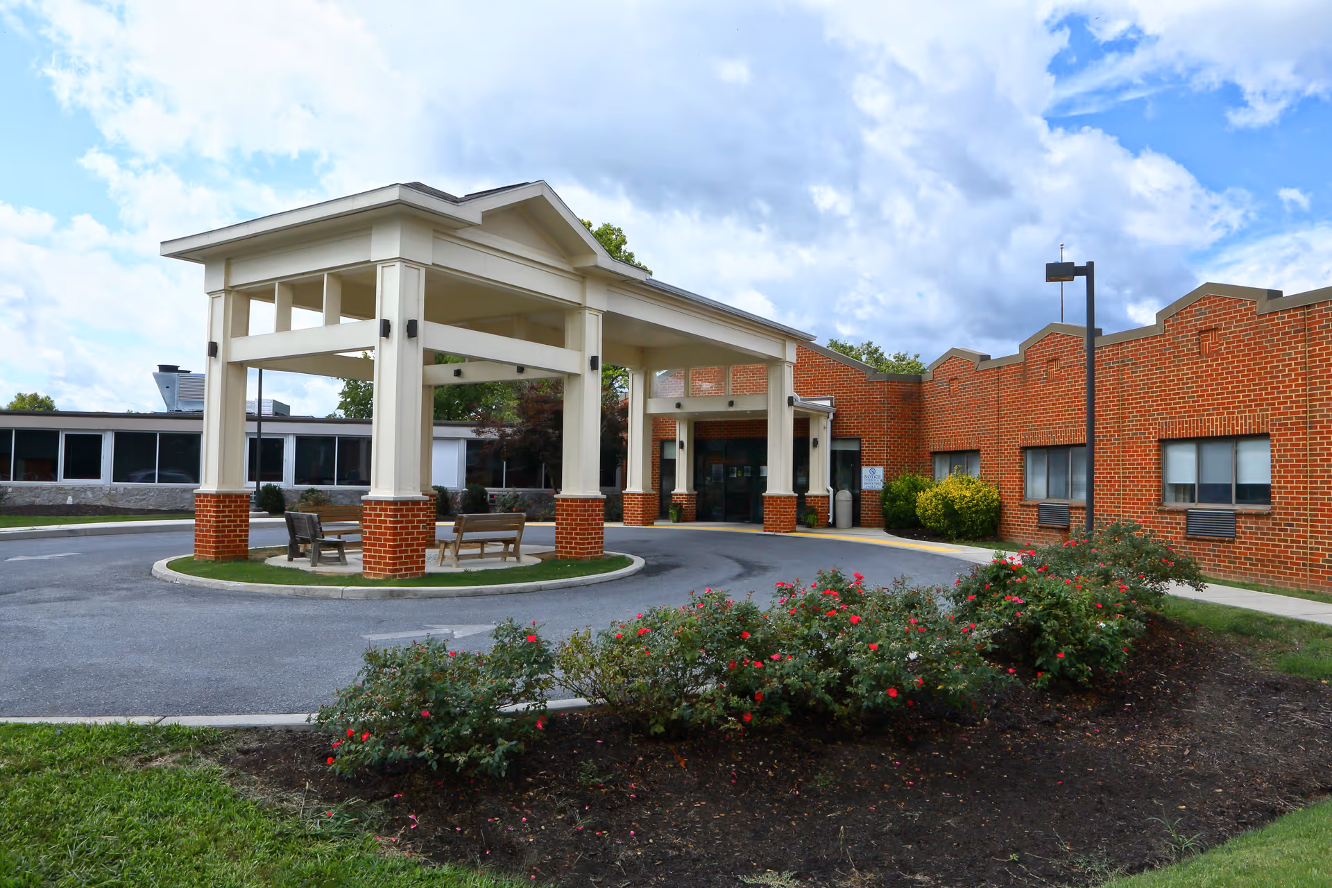 Exterior view of a senior living facility with a covered entrance supported by white columns with brick bases. There are benches under the covered area and a circular driveway. The building is made of red brick with several windows, and there are bushes with red flowers in the foreground under a partly cloudy sky.