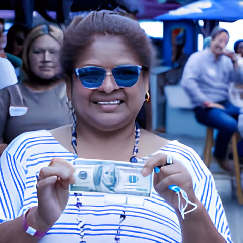 A smiling woman wearing blue sunglasses and a striped shirt holds up a folded dollar bill. She is outdoors with other people sitting and standing in the background under blue umbrellas.