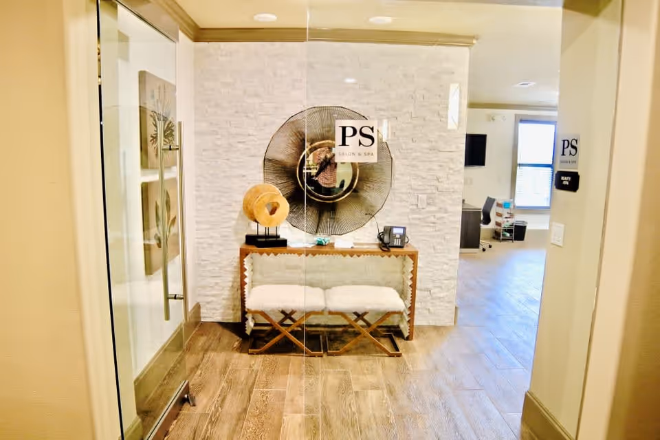 Interior view of a salon and spa reception area with a wooden console table, decorative items, and two cushioned stools. The wall behind the table has a large circular decorative piece and a sign that reads 'PS Salon & Spa'. The floor is wooden, and there is a glass door on the left side.