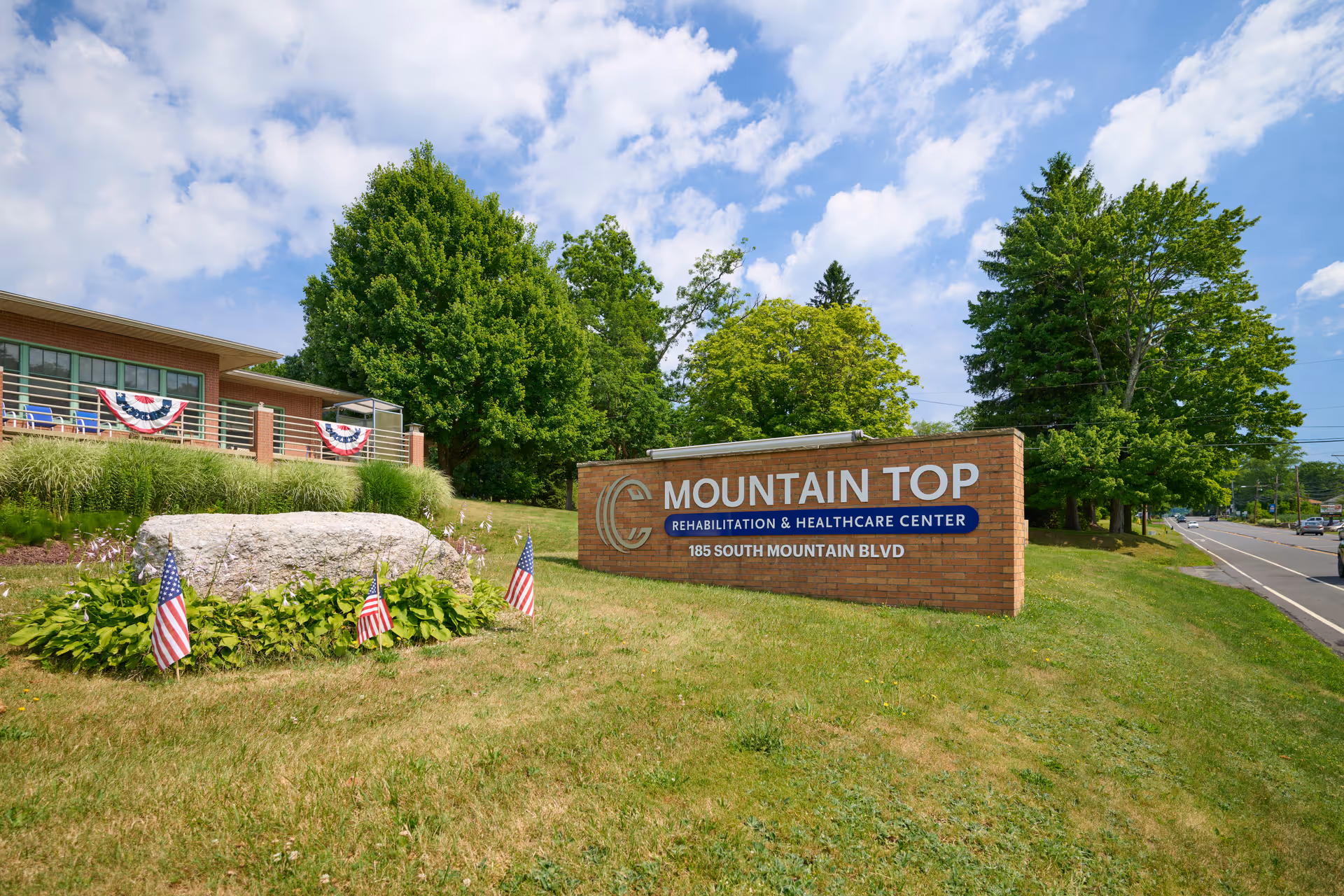Outdoor view of Mountain Top Rehabilitation and Healthcare Center sign on a grassy area with small American flags and a large rock. The building is partially visible in the background with patriotic bunting on the railing. Trees and a road are also visible under a partly cloudy sky.