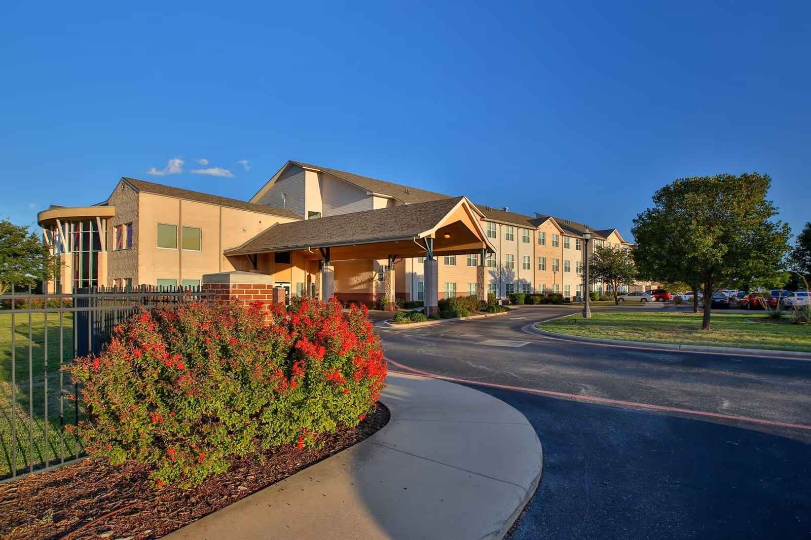Exterior view of Wesley Court senior living facility showing a large multi-story building with a covered entrance, surrounded by landscaped bushes with red flowers, a tree, and a parking area under a clear blue sky.