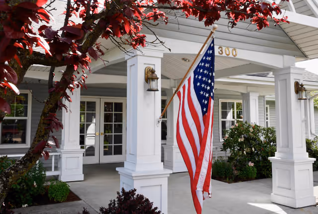 Entrance of a senior living facility with white pillars, an American flag hanging from one pillar, and the number 300 displayed above the doorway. There are plants and bushes around the entrance and a tree with red leaves in the foreground.