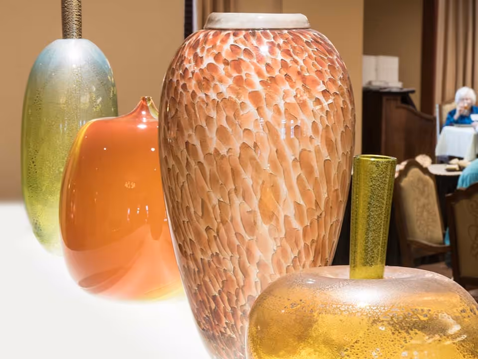 Close-up view of four decorative glass vases in various colors and shapes displayed on a white surface, with a blurred background showing elderly people seated at tables in a common area.