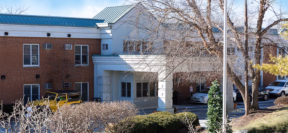 Exterior view of a brick building with a covered entrance and several windows. There are leafless trees and bushes in the foreground, along with parked cars near the entrance.