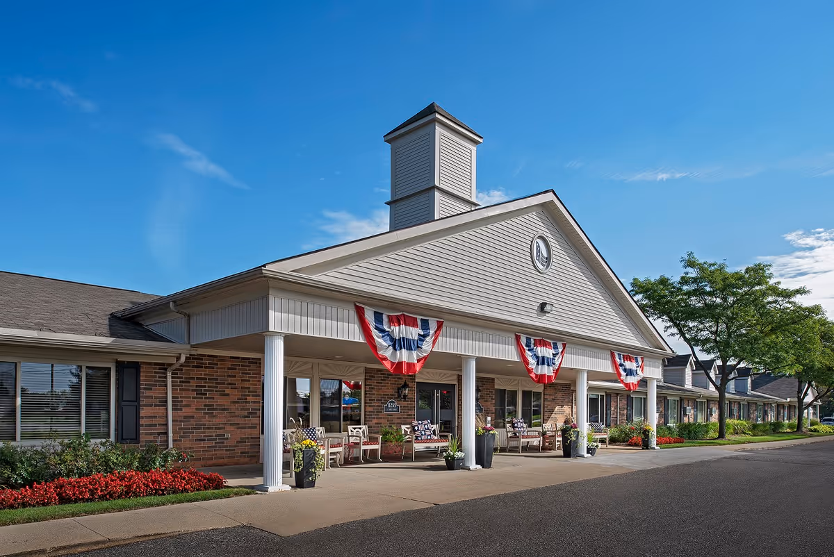 Exterior view of American House Sterling Heights, a single-story brick building with white columns and a covered entrance decorated with red, white, and blue bunting. There are benches and potted plants near the entrance, with a clear blue sky and trees in the background.
