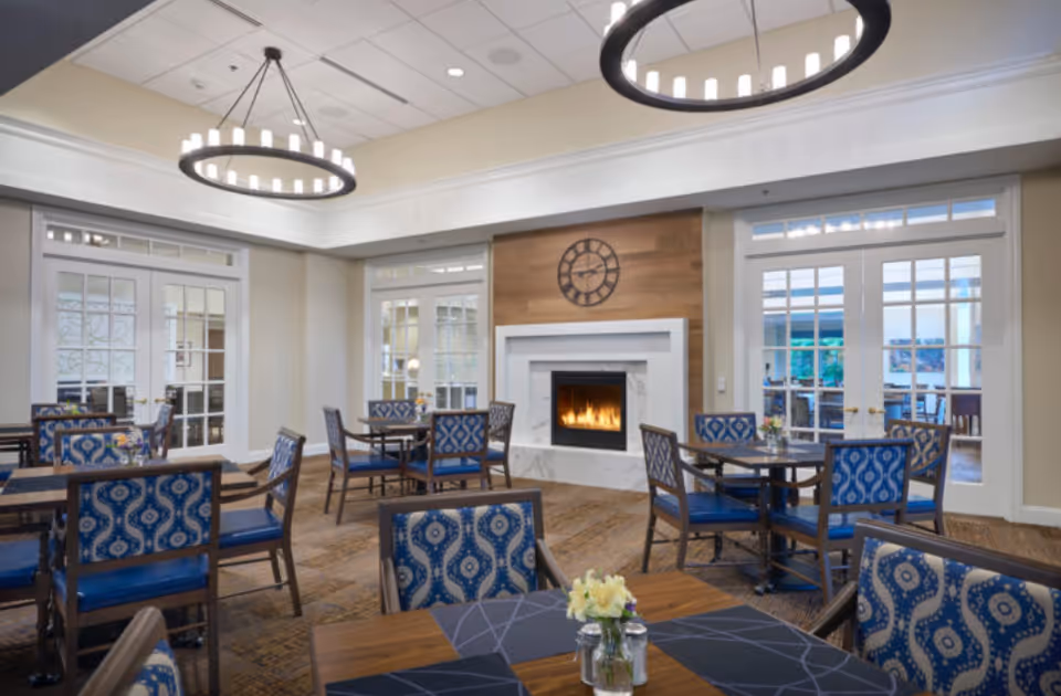 A cozy dining room with multiple tables and blue patterned chairs. The room features a lit fireplace with a wooden accent wall and a large clock above it. Two modern circular chandeliers hang from the ceiling, and there are large glass doors and windows letting in natural light.