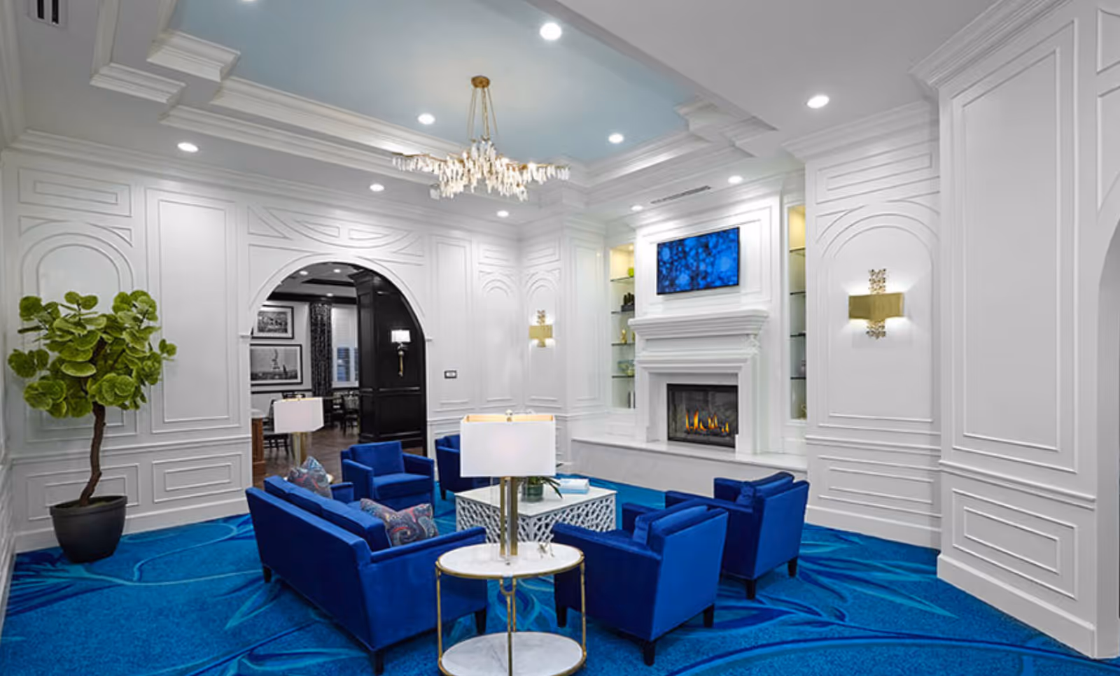 A luxurious living room with white paneled walls and ceiling, a blue patterned carpet, and blue upholstered sofas and armchairs arranged around a white coffee table. A modern chandelier hangs from the ceiling, and a fireplace with a mounted flat-screen TV above it is on one wall. A potted plant is in the corner, and there is an arched doorway leading to another room.