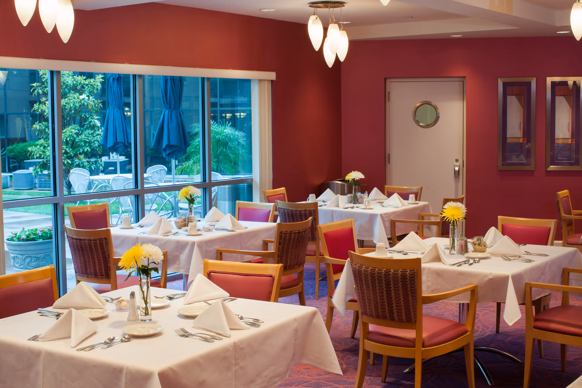 A dining room with several tables covered in white tablecloths, each set with folded white napkins, plates, silverware, and small flower vases. The room has red walls and large windows showing an outdoor patio area with chairs and umbrellas.