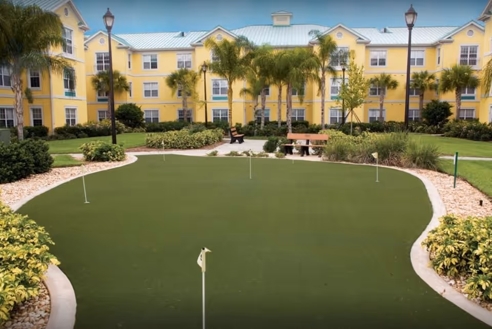 Outdoor putting green with several small flags, surrounded by landscaping with bushes and palm trees, benches, and a yellow three-story building in the background under a blue sky.