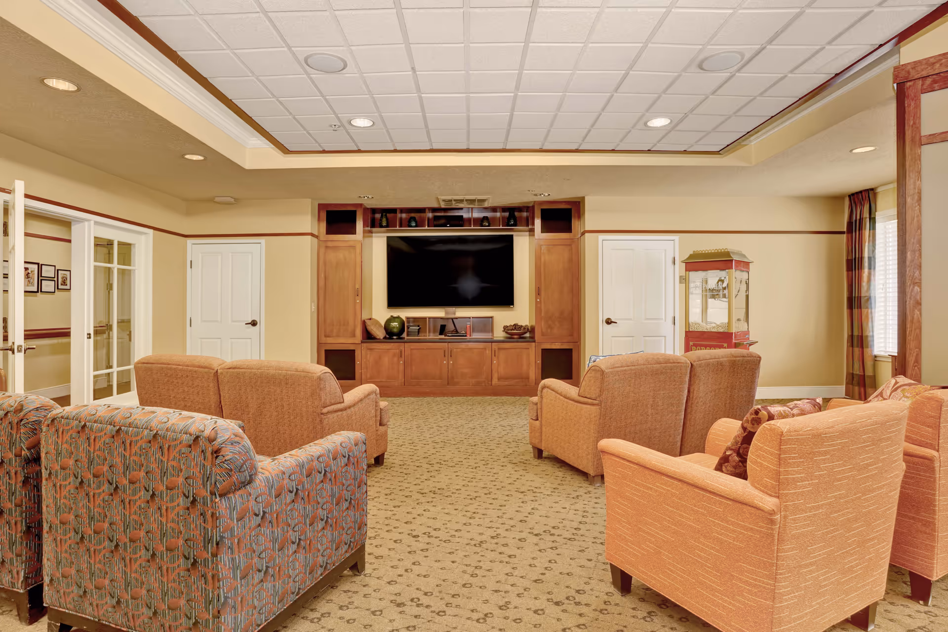 A cozy living room area with multiple upholstered armchairs and sofas arranged facing a large flat-screen TV mounted on a wooden entertainment center. The room has beige walls, a patterned carpet, recessed lighting in the ceiling, and a popcorn machine in the corner near a window with plaid curtains.