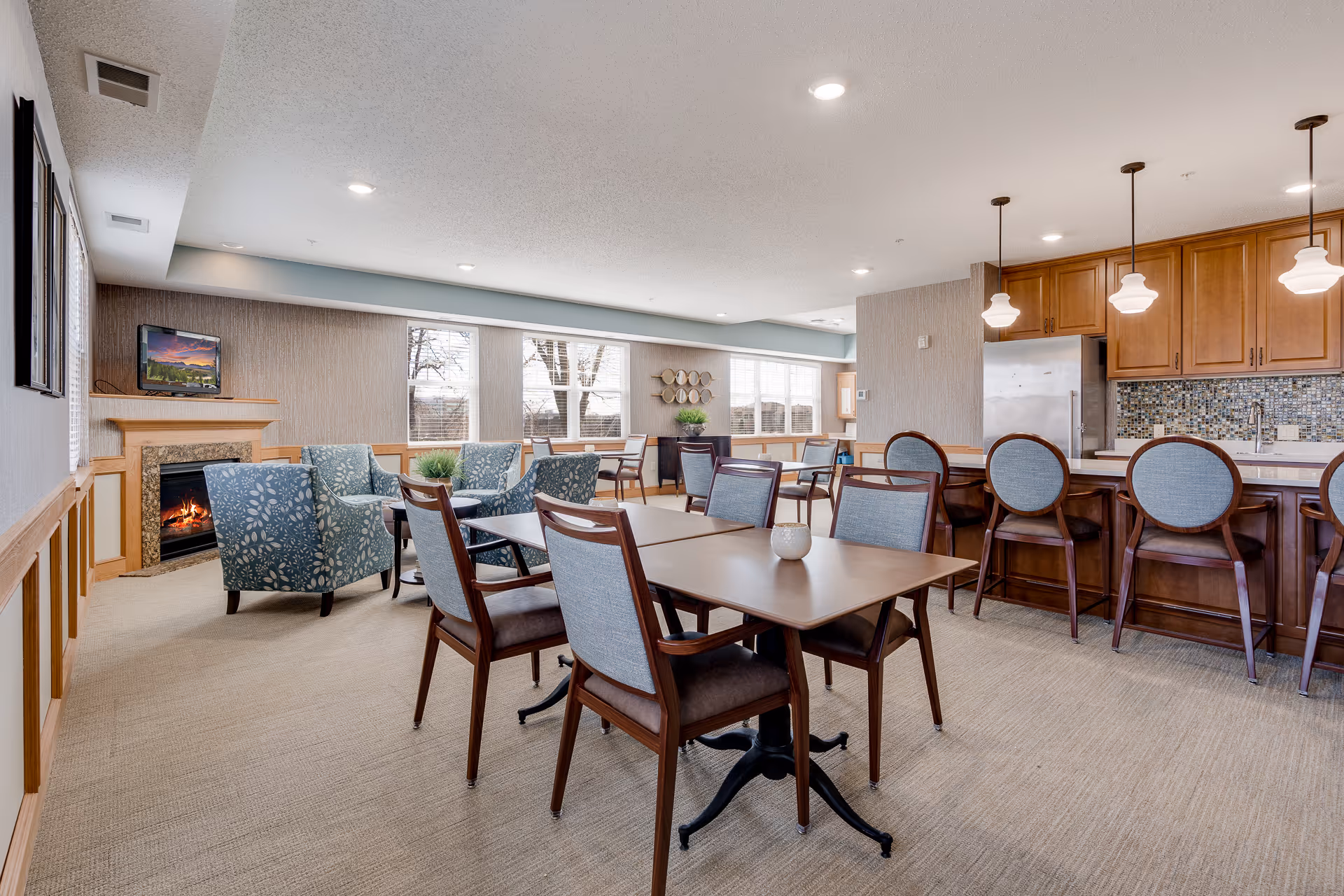 A bright and spacious common area in Cherrywood Pointe Assisted living facility featuring a cozy seating area with patterned armchairs around a fireplace, multiple dining tables with chairs, and a kitchen area with a counter and bar stools under pendant lights. Large windows let in natural light, and the room has a warm, inviting atmosphere.