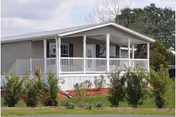 Single-story building with a covered front porch featuring white railings and columns, surrounded by small bushes and grass under a cloudy sky.