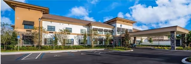 Exterior view of Bella Mar Royal Palm Beach facility showing a two-story building with a covered entrance, surrounded by landscaped greenery and a parking lot with handicap parking spaces under a partly cloudy sky.