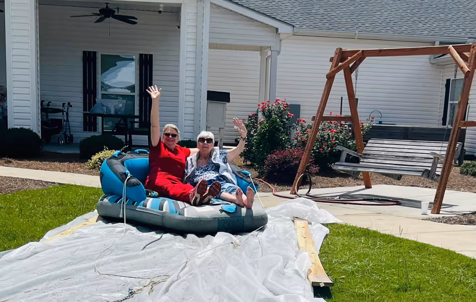 Two women sitting on an inflatable raft on a plastic sheet outdoors in a grassy area. One woman is wearing a red outfit and sunglasses, and the other is wearing a patterned top and sunglasses. Both are smiling and waving. In the background, there is a white building with a porch, some bushes with red flowers, and a wooden swing.