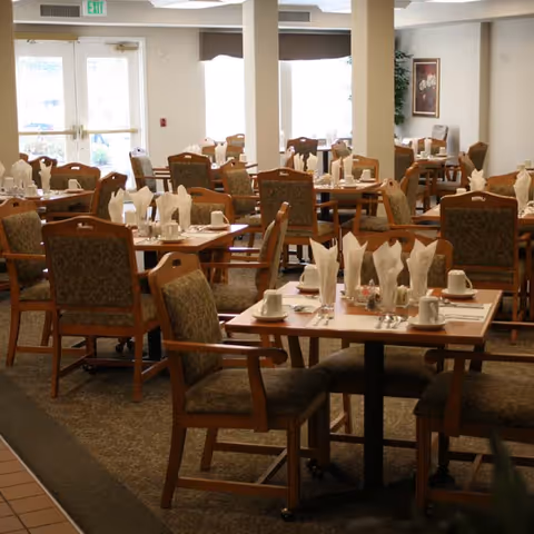 A dining room with multiple wooden tables and cushioned chairs arranged neatly. Each table is set with white napkins folded upright, cups, saucers, and silverware. Large windows in the background allow natural light to fill the room, and there is a framed picture and a potted plant on the far wall.