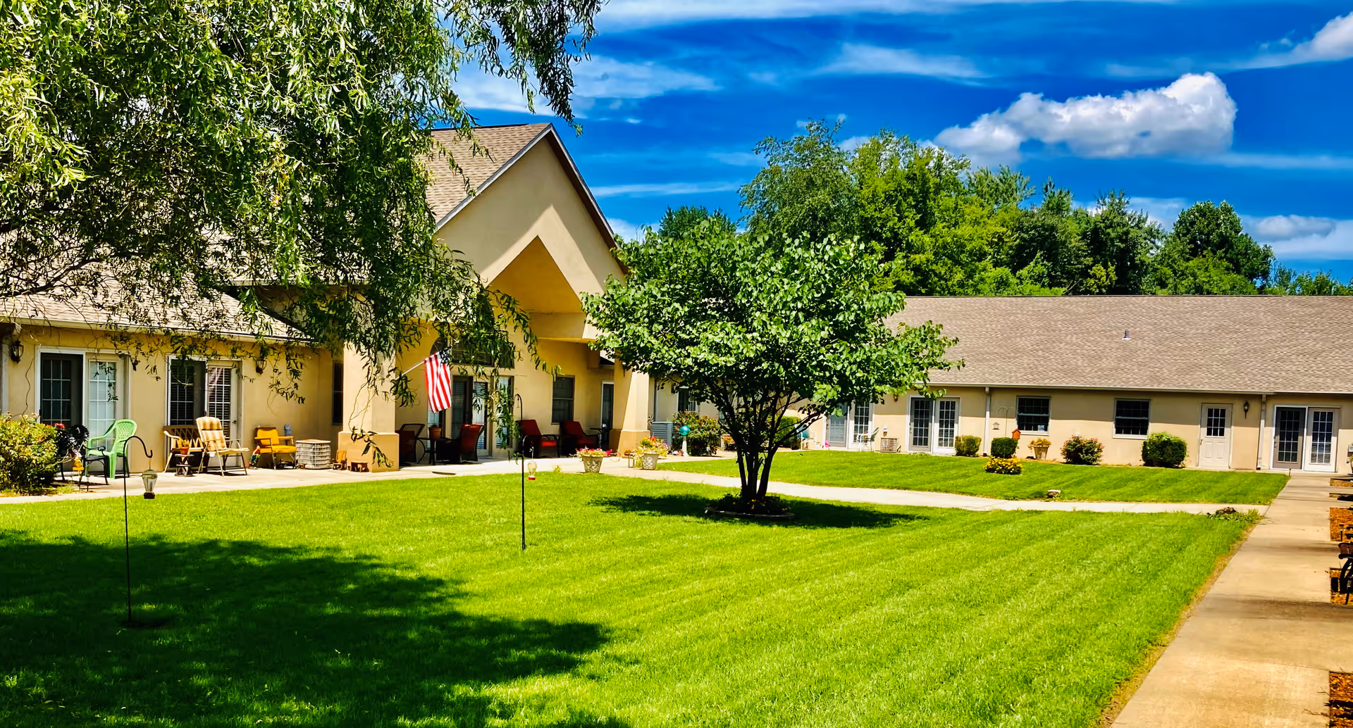 A sunny outdoor courtyard at Southgate Senior Living featuring a well-maintained green lawn, a central tree, and a beige building with multiple doors and windows surrounding the courtyard. There are chairs and potted plants along the building's walkway, and an American flag is displayed near the entrance.