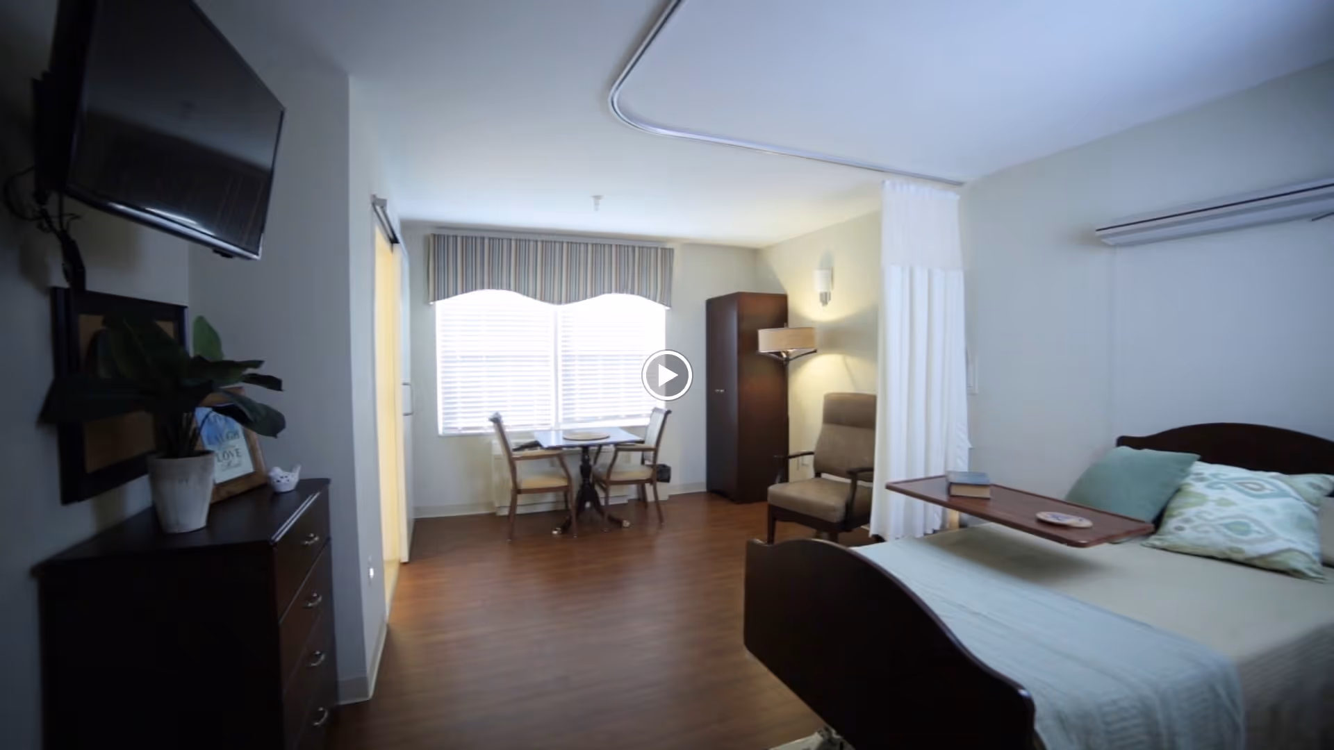 A well-lit senior living bedroom with a bed featuring pillows and a bedside tray, a comfortable armchair, a small dining table with two chairs near a window with blinds and a valance, a tall cabinet, a dresser with a plant and framed picture, and a wall-mounted TV.