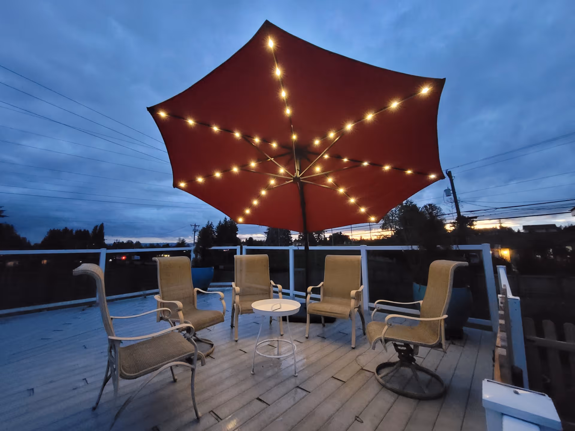 Outdoor patio area at dusk with six beige cushioned chairs arranged around a small white round table. A large red umbrella with string lights is open above the seating area. The sky is cloudy and darkening, with some trees and power lines visible in the background.