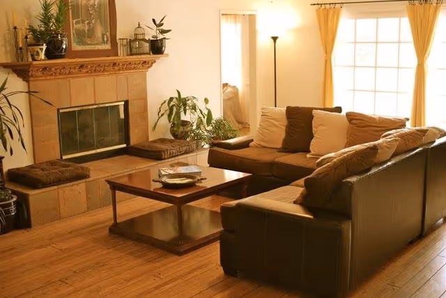 A cozy living room with a brown sectional sofa adorned with beige and brown cushions, a wooden coffee table with decorative items, a fireplace with a wooden mantle decorated with plants and framed artwork, and large windows with yellow curtains allowing natural light to fill the room.