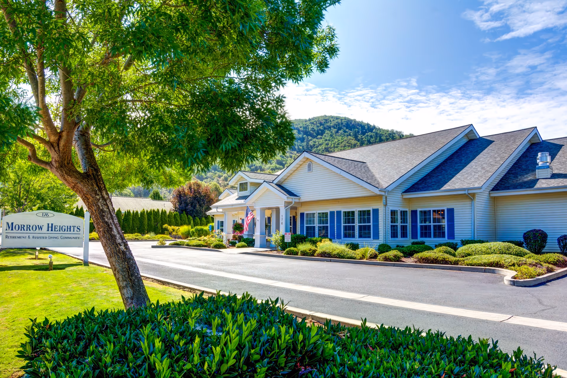 Exterior view of Morrow Heights Assisted Living facility with a well-maintained driveway, landscaped bushes, a large tree, and a sign that reads 'Morrow Heights Retirement & Assisted Living Community' under a bright blue sky with some clouds and a mountain in the background.