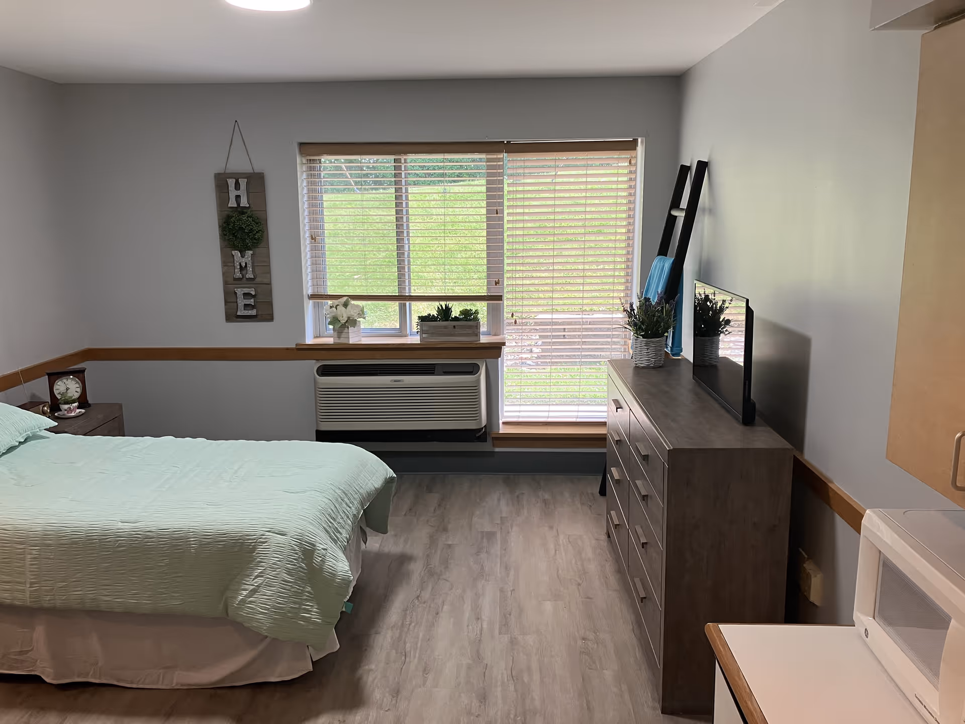 A neat and cozy bedroom with a bed covered in a light green quilt, a nightstand with a clock and teacup, a window with wooden blinds letting in natural light, a dresser with a flat-screen TV and potted plants, a decorative ladder with a blue towel, and a microwave on a counter to the right.