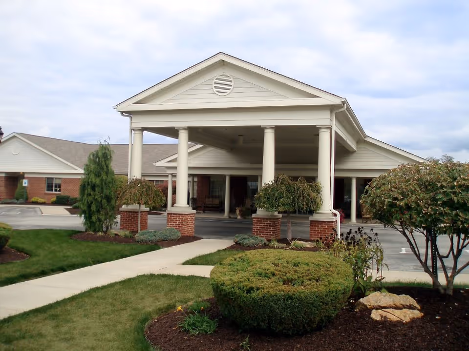 Front exterior view of North Strabane Retirement Village showing a covered entrance with white columns, brick accents, landscaped bushes, and a walkway leading to the entrance under a cloudy sky.