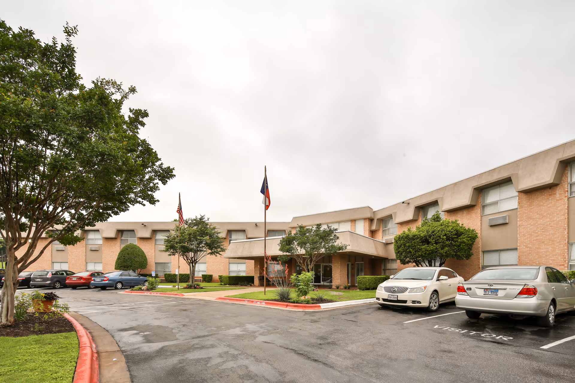 Exterior view of Heritage Park Rehabilitation and Skilled Nursing Center showing a two-story brick building with multiple windows, a covered entrance, several parked cars, and landscaped greenery including trees and bushes under an overcast sky.
