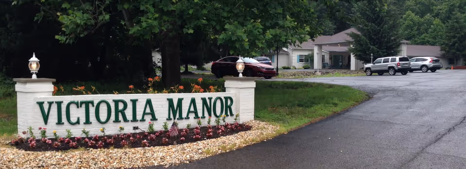 Entrance sign for Victoria Manor with green lettering on a white brick structure surrounded by flowers and two lamp posts, with a driveway leading to a building and parked cars in the background.