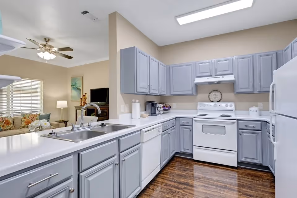 A clean and modern kitchen with light gray cabinets, white countertops, and white appliances including a stove, refrigerator, and dishwasher. The kitchen has a double sink with a chrome faucet and overlooks a living room area with a ceiling fan, a couch with colorful pillows, a lamp, and a TV on a stand.
