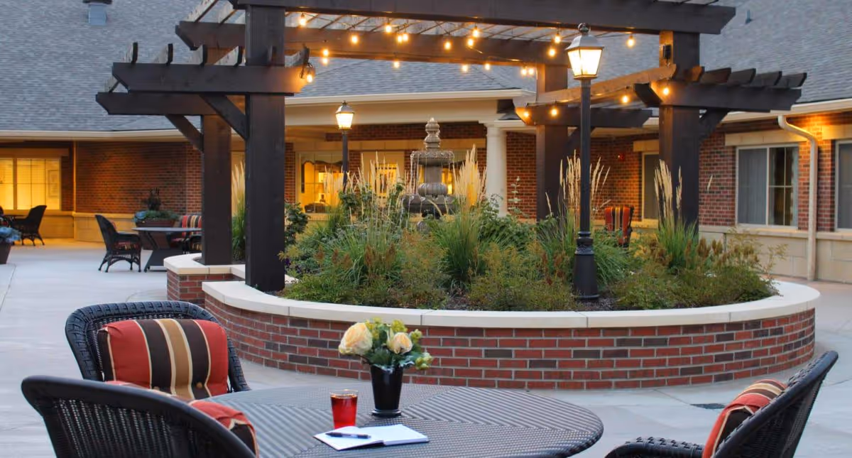 Outdoor courtyard area at Deer Ridge Memory Care featuring a round table with chairs that have red and striped cushions, a small vase with flowers, and a glass of red drink. In the background, there is a raised circular garden bed with ornamental grasses and a multi-tiered water fountain under a wooden pergola with string lights. The building's brick exterior and windows are visible, along with lampposts providing warm lighting.