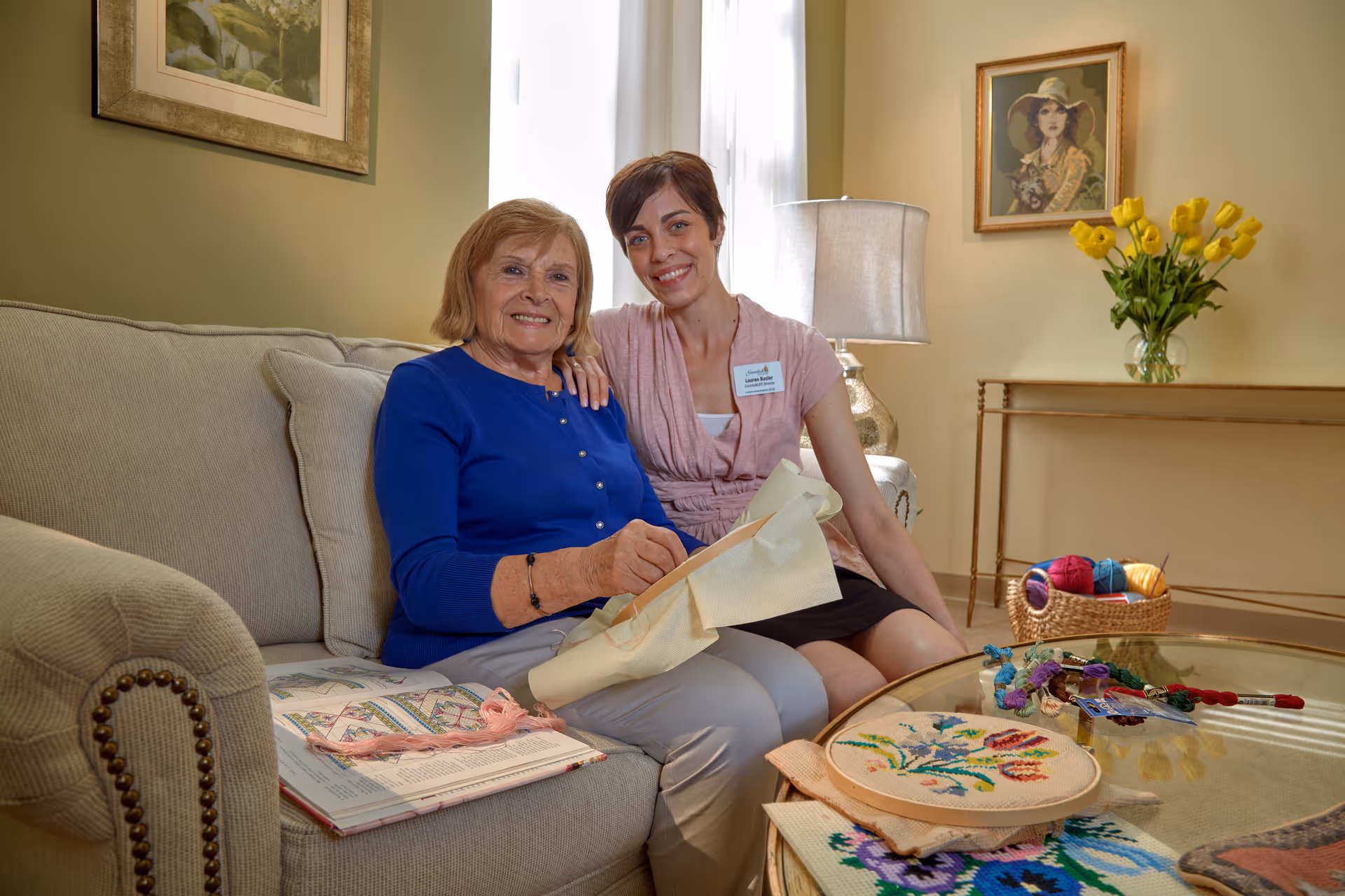 An elderly woman and a younger woman sitting together on a beige couch in a cozy living room. The elderly woman is holding an embroidery hoop with a floral design, and there are embroidery supplies on the glass coffee table in front of them. The younger woman, wearing a name tag, has her arm around the elderly woman and they are both smiling. The room has light-colored walls, framed artwork, a table lamp, a vase with yellow flowers, and a basket with colorful yarn balls.