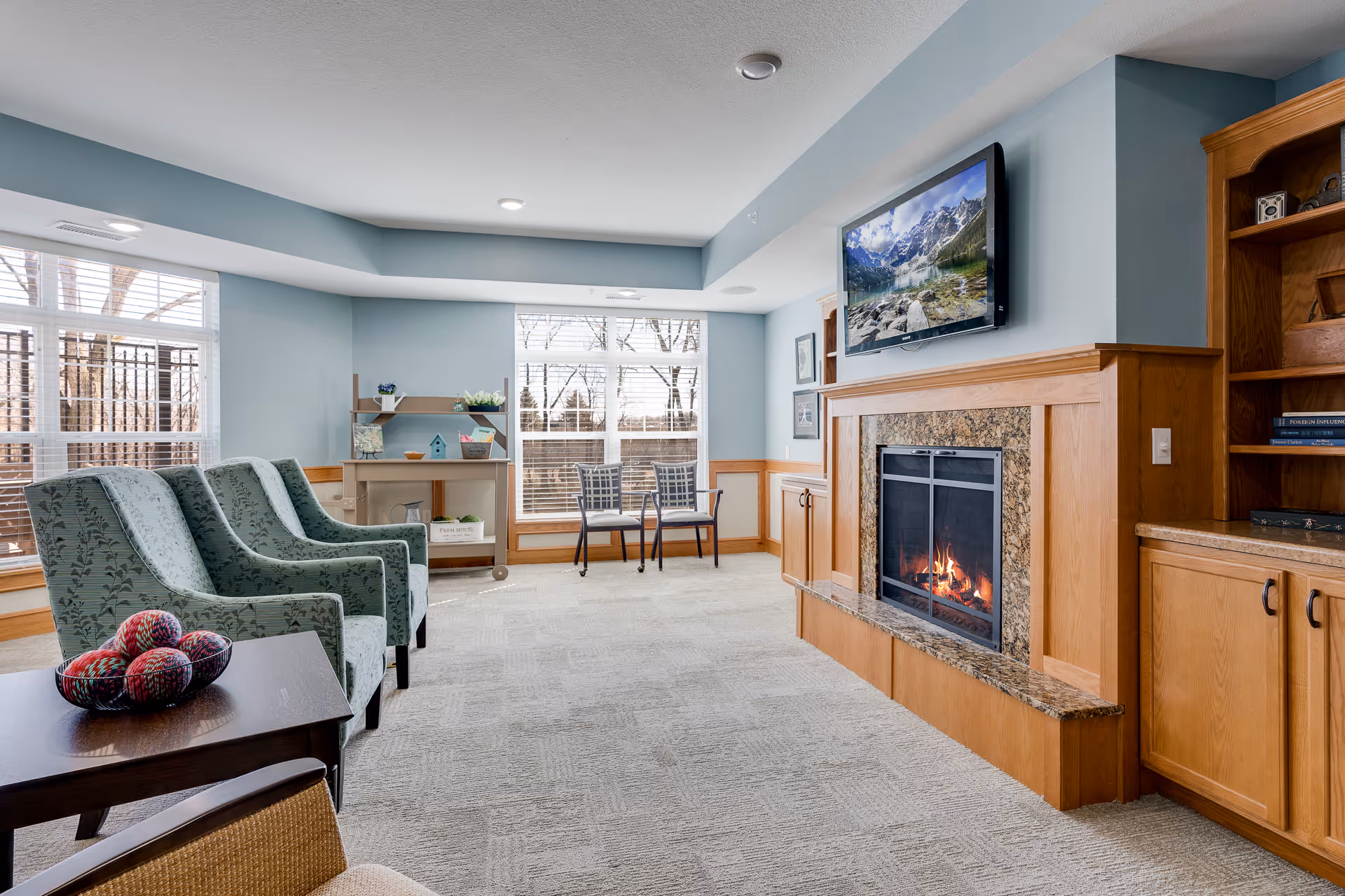 A cozy living room area in Cherrywood Pointe Assisted featuring a lit fireplace with a granite surround and wooden mantel. Above the fireplace is a flat-screen TV displaying a mountain lake scene. The room has light blue walls with wood paneling, large windows with white blinds letting in natural light, and several chairs including two patterned armchairs and two metal chairs near the windows. There is a small table with decorative yarn balls and a wooden shelving unit with books and decor items.