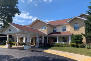 Exterior view of a two-story senior living facility building with beige brick walls and a red roof. The entrance has a covered driveway supported by white columns. There are green shrubs and flowers along the front of the building, and a clear blue sky with some clouds above.