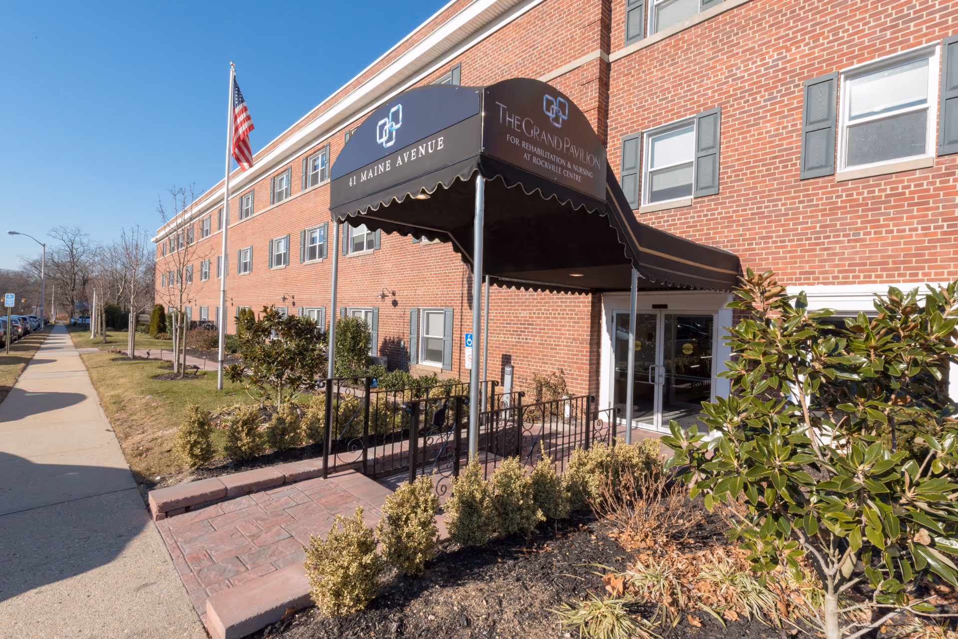 Exterior view of The Grand Pavilion for Rehabilitation and Nursing at Rockville Centre, showing a brick building with multiple windows, a black awning over the entrance with the facility's name and address, a flagpole with an American flag, and landscaped bushes and plants along the walkway.