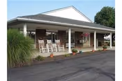 Front exterior view of Alexandria Care Center showing a covered porch with white rocking chairs, potted plants, and a paved driveway. The building has a brick facade and a white roof trim.