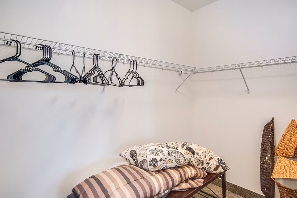 A small room with white walls featuring wire shelving along two walls with several black plastic hangers. Below the shelves is a bench with folded bedding including a striped blanket and a floral pillow. Two woven baskets are placed on the floor in the corner.