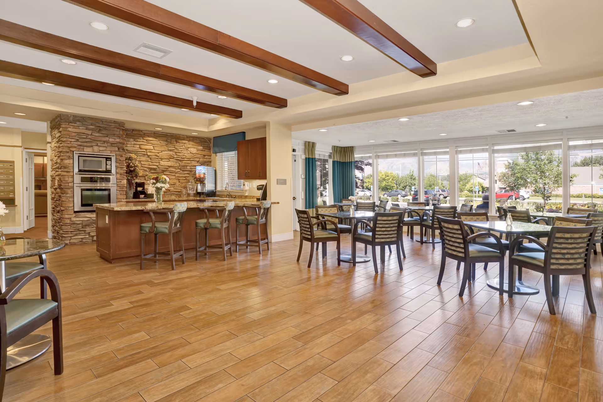 A spacious and bright dining area with multiple tables and chairs arranged neatly. The room features large windows allowing natural light to fill the space and providing a view of the outdoors. There is a kitchen area with a stone accent wall, built-in ovens, a countertop with bar stools, and cabinetry. The floor is wooden, and the ceiling has recessed lighting with wooden beams.