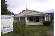 Single-story white building with a covered porch and an American flag in front, surrounded by greenery and bushes. A sign in the foreground reads 'Evergreen Manor Skilled Nursing and Rehabilitation Center.'