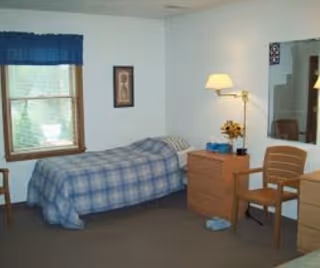 A simple bedroom with a single bed covered in a blue plaid blanket, a wooden nightstand with a lamp and a small plant, a wooden chair, a window with a blue valance, and a framed picture on the wall.