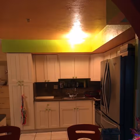 Interior view of a kitchen with white cabinets, a stainless steel refrigerator, a sink, and a countertop. The ceiling has a yellow-green accent with a bright ceiling light. There are some kitchen items on the counter and a red chair partially visible in the foreground.