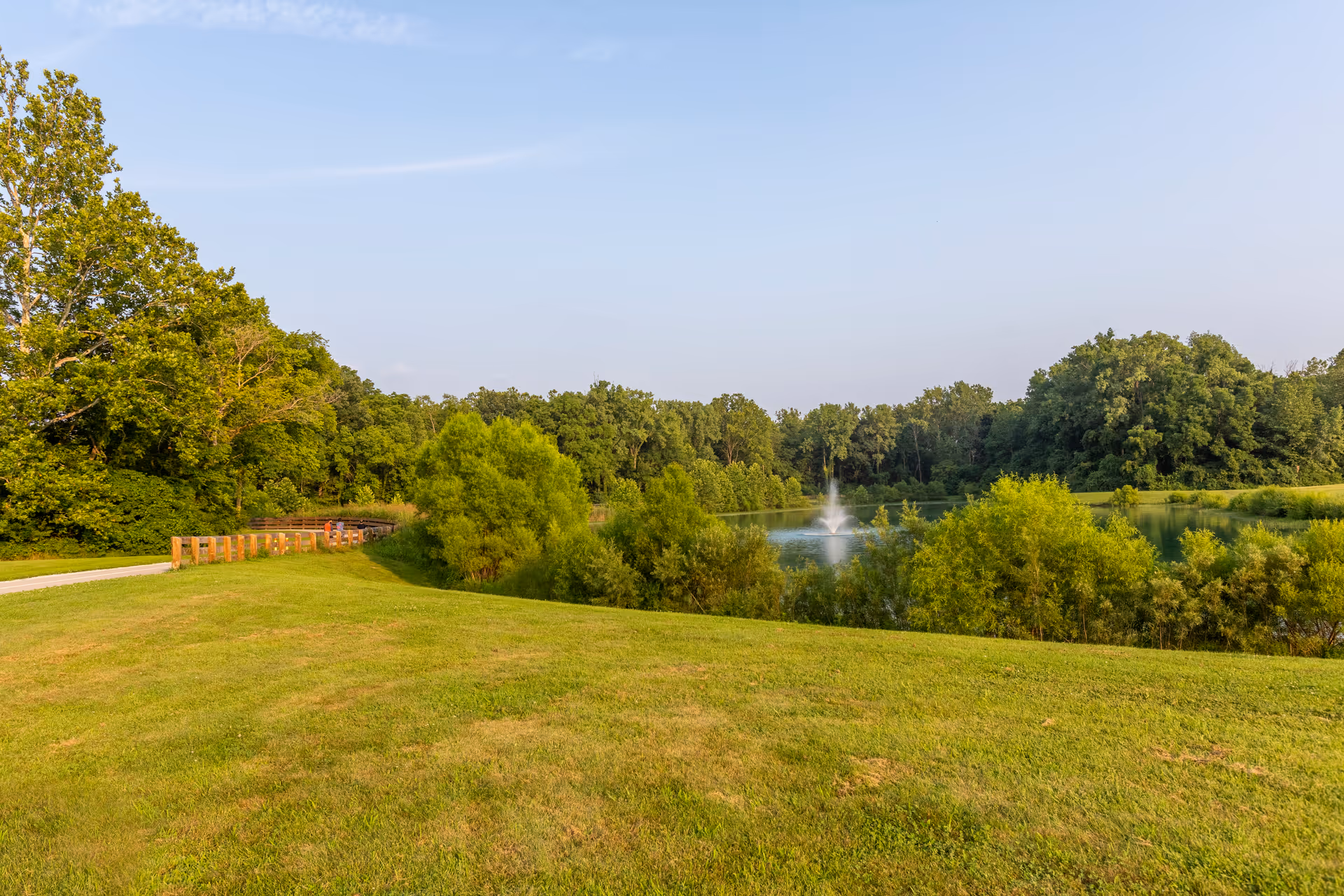 A scenic outdoor view featuring a grassy area with a walking path on the left, surrounded by lush green trees. In the background, there is a pond with a water fountain spraying water upwards under a clear blue sky.