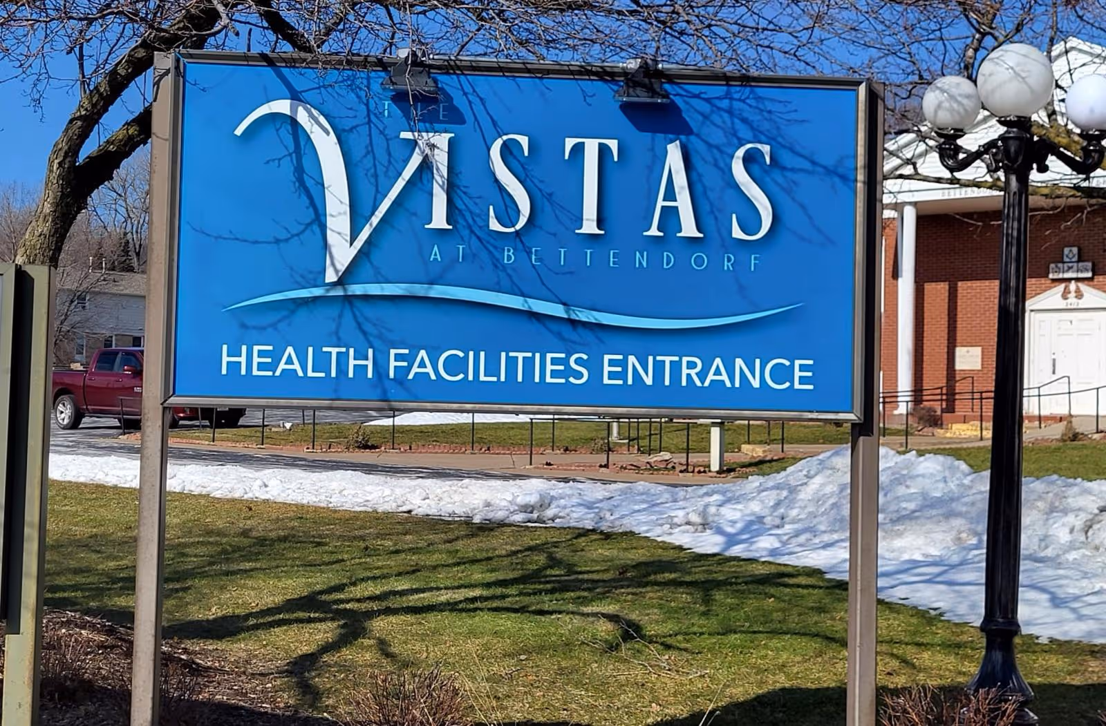 Outdoor image of a blue sign for The Vistas at Bettendorf health facilities entrance, with a building and lamp post visible in the background and patches of snow on the grass.