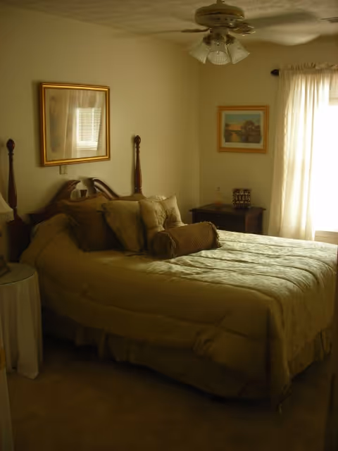 A cozy bedroom with a neatly made bed featuring multiple pillows and a bolster. The room has beige walls, a ceiling fan with light fixtures, framed artwork on the walls, a window with sheer curtains allowing natural light to enter, and a small bedside table with decorative items.