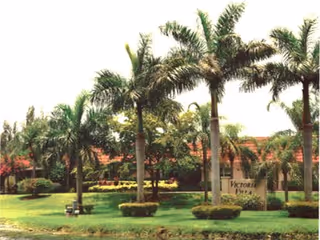 Outdoor view of Victoria Villa facility with several tall palm trees, green grass, and landscaped bushes in front of a building with a red roof. A sign reading 'Victoria Villa' is visible among the trees.